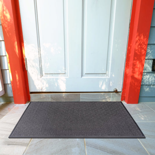 A rectangular, charcoal gray indoor/outdoor entrance mat made of polypropylene with a ribbed texture, placed on a tiled floor in front of a white door with red trim.