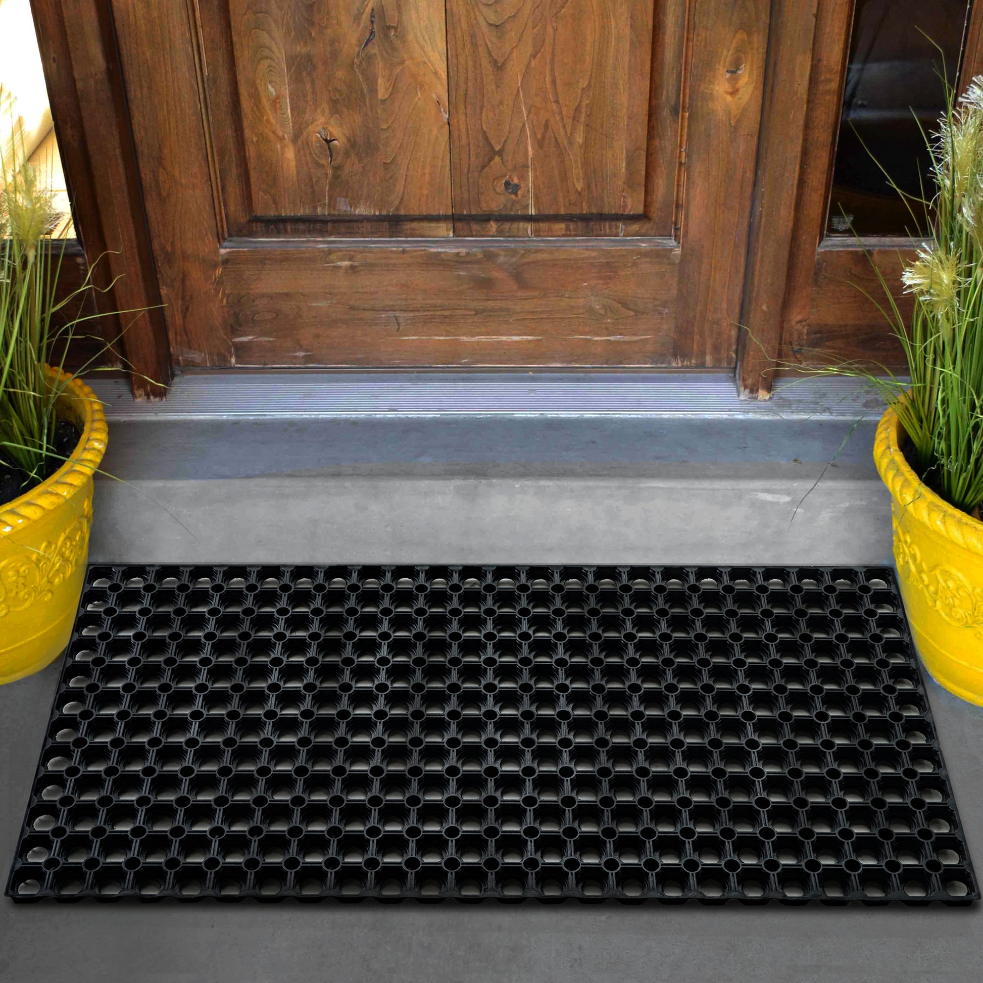 A black Hometex All Seasons Outdoor Entrance Mat placed in front of a wooden door with yellow potted plants on either side.
