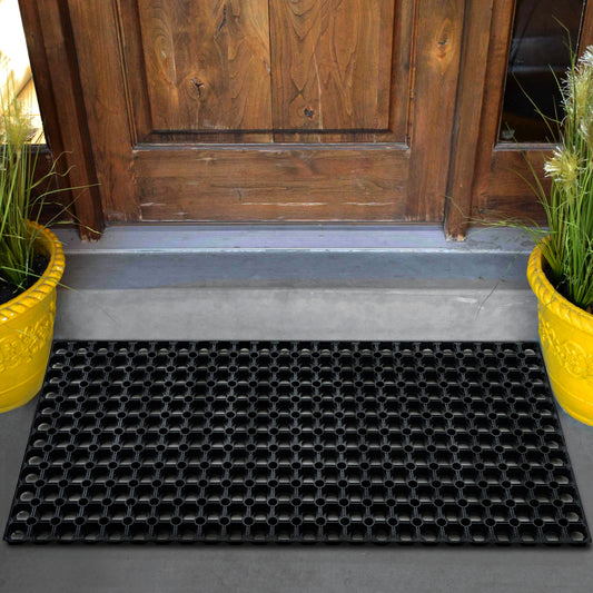 A black Hometex All Seasons Outdoor Entrance Mat placed in front of a wooden door with yellow potted plants on either side.